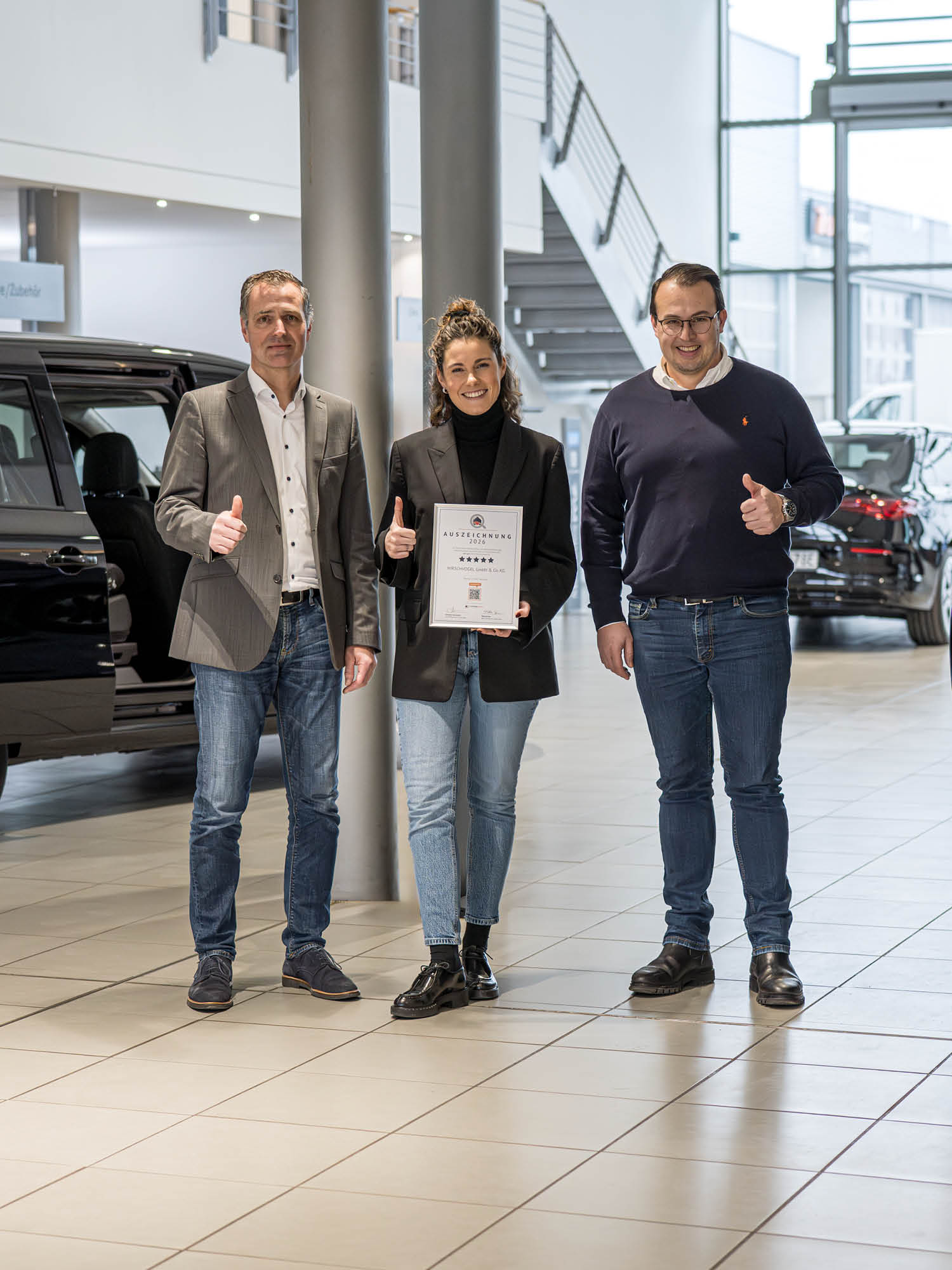 In einem großzügigen, hellen Autohaus stehen drei Personen nebeneinander. Alle drei zeigen mit einer Hand ein „Daumen hoch“-Zeichen. Die Person in der Mitte hält eine Urkunde vor dem Körper. Hinter der Gruppe sind mehrere Fahrzeuge sowie große Fensterfronten und architektonische Elemente des Autohauses sichtbar. Die Szene vermittelt eine feierliche und wertschätzende Übergabe‑ oder Auszeichnungssituation.