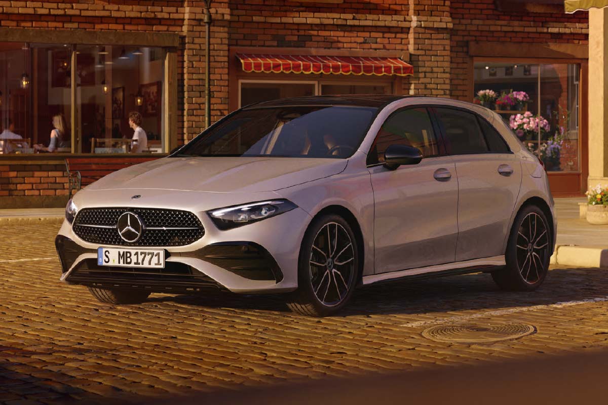 Das Bild zeigt ein helles, modernes Fahrzeug der Marke Mercedes‑Benz, das auf einer gepflasterten Straße in einer städtischen Umgebung steht. Das Auto befindet sich leicht schräg zur Kamera, sodass sowohl die Front als auch die Seite sichtbar sind. Die Frontpartie weist einen großen Kühlergrill mit Mercedes‑Benz‑Logo sowie schmale Scheinwerfer auf. Das Fahrzeug hat fünf Türen und schwarze Felgen.