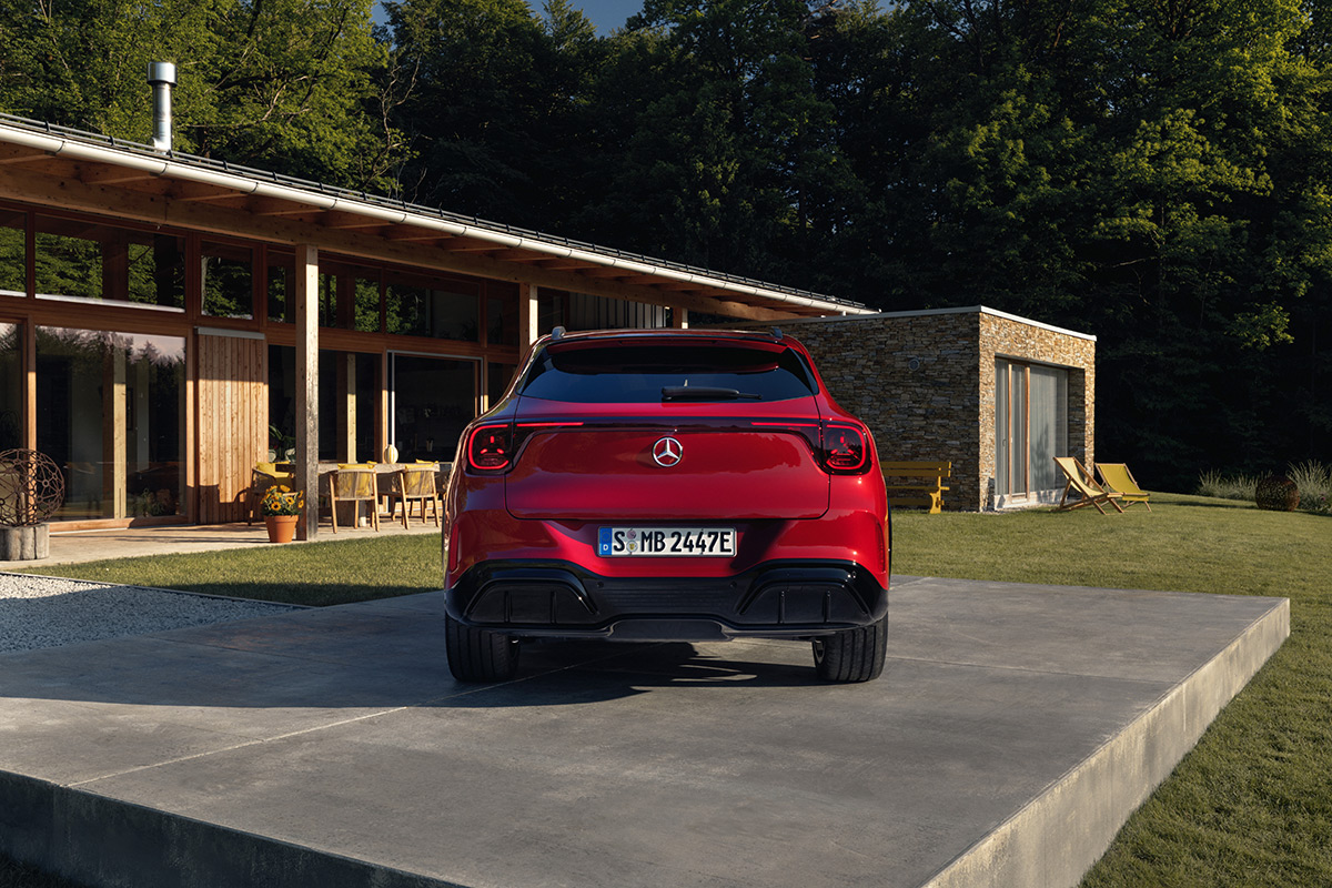 Heckansicht eines roten Mercedes SUV auf einer Betonflaeche vor einem modernen Wohnhaus mit grosser Fensterfront in naturnaher Umgebung.