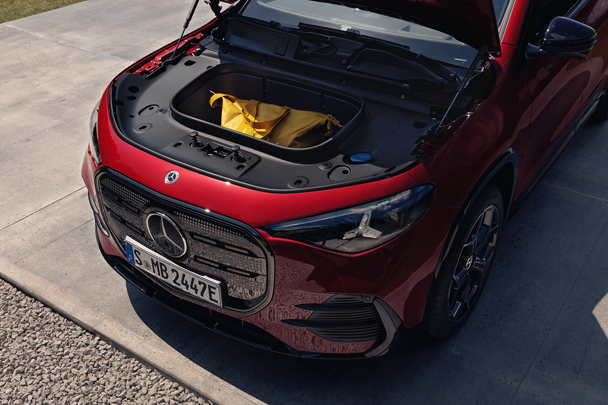 Geoeffnete Fronthaube eines roten Mercedes Elektro SUV mit vorderem Stauraum und Tasche vor einem modernen Wohngebaeude.