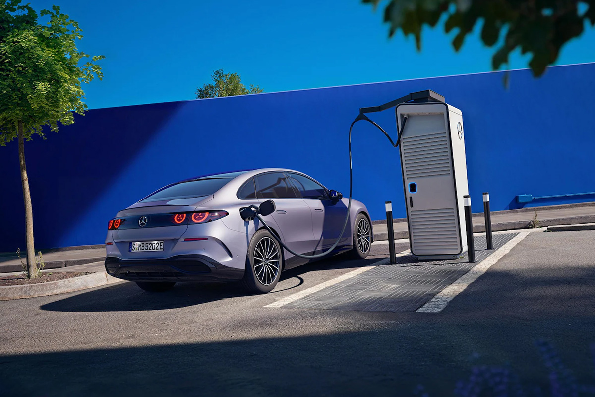 Heckansicht einer silbergrauen Mercedes Benz C Klasse an einer Ladestation. Das Elektroauto ist ueber ein Ladekabel mit der Ladesaeule verbunden. Im Hintergrund eine markante blaue Wand unter blauem Himmel.