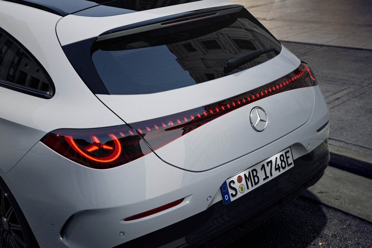 Heckansicht eines weissen Mercedes Kombis mit durchgehendem LED-Ruecklicht und leuchtenden Rueckleuchten, stehend auf einer Strasse in einer Stadtumgebung.