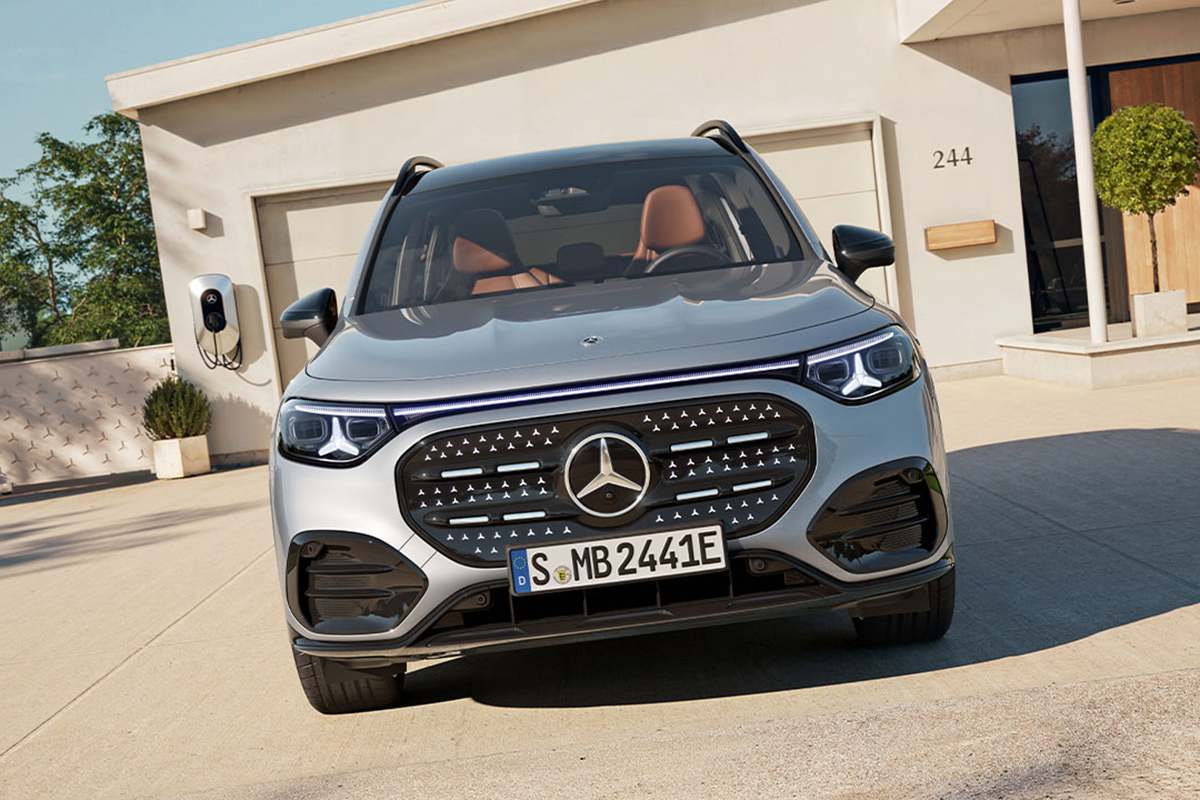 Frontansicht eines silbernen SUV in einer sonnigen Einfahrt. Sichtbar sind LED‑Scheinwerfer, markanter Grill mit Muster, schwarze Felgen und ein Ladeanschluss an der Hauswand im Hintergrund.