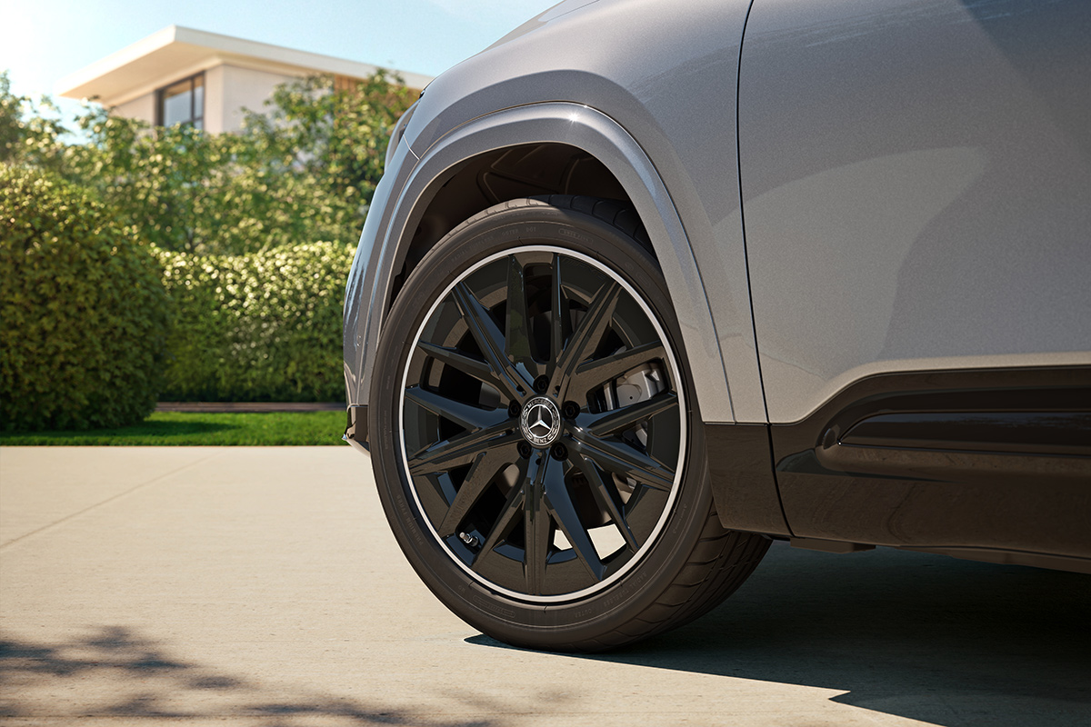 Nahaufnahme eines schwarzen Mercedes Leichtmetallrads an einem silbergrauen SUV, aufgenommen bei Tageslicht vor gruener Bepflanzung und einem modernen Haus im Hintergrund.