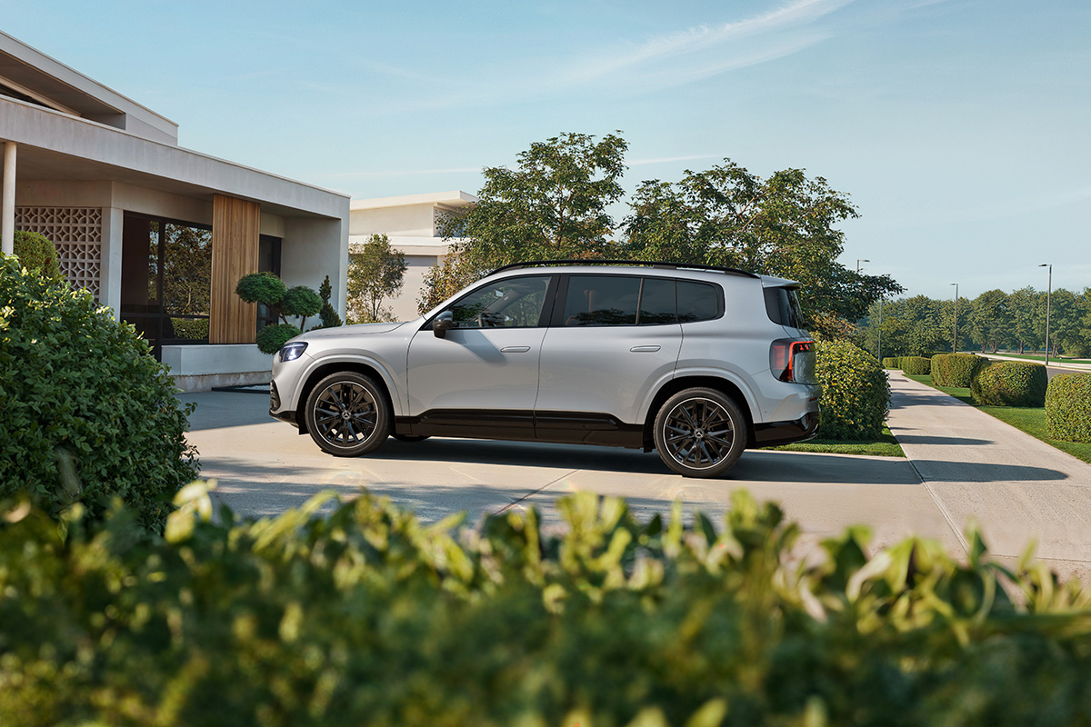 Seitliche Ansicht eines silbernen Mercedes SUV, geparkt vor einem modernen Haus, umgeben von gruener Bepflanzung und Bueschen bei klarem Tageslicht.