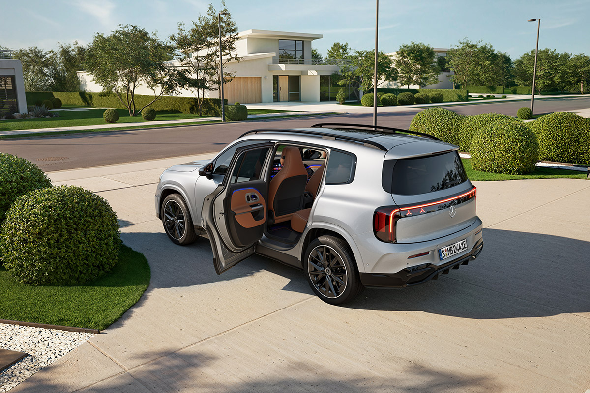 Silberner SUV steht in einer modernen Einfahrt mit geoeffneter Fondtuere. Sichtbar sind schwarze Felgen, braune Innenverkleidung, klare Karosserielinien und umgebende gruene Bepflanzung.