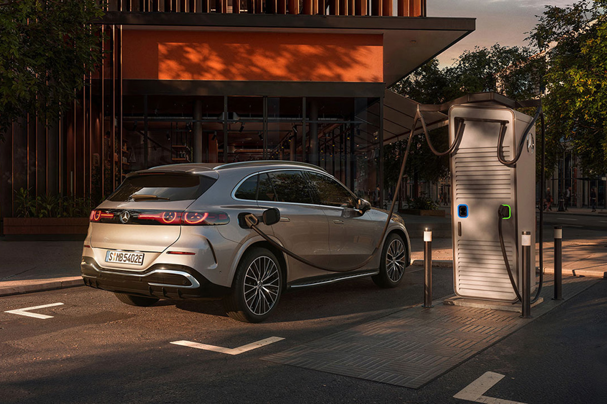 Silberner SUV steht an einer Ladestation in einer stadtlichen Umgebung. Sichtbar sind LED‑Rueckleuchten, schwarze Felgen, das angeschlossene Ladekabel und die moderne Architektur im Hintergrund.