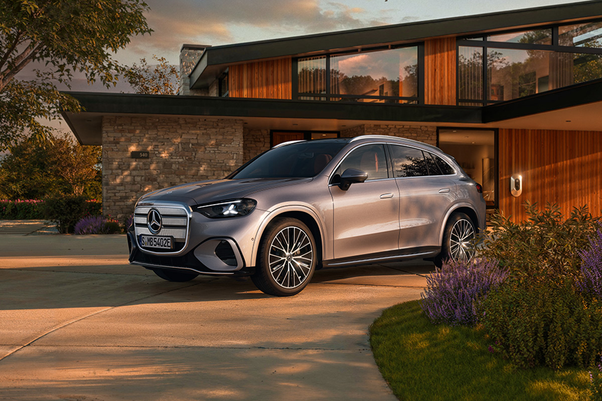 Silberner Mercedes-Benz GLC EV geparkt auf einer Auffahrt vor einem modernen Wohnhaus mit Holzfassade im warmen Abendlicht. Im Vordergrund bluehende Lavendelstraeucher