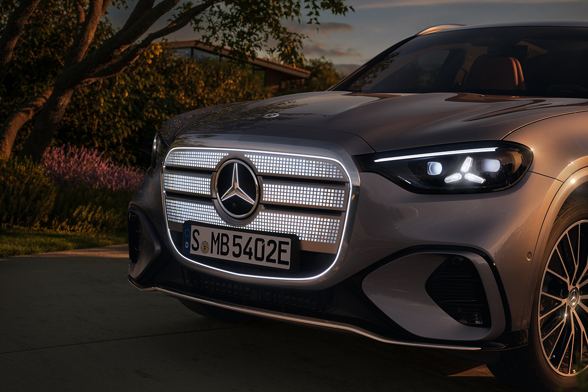 Frontansicht eines silbergrauen Mercedes Elektro-SUV mit leuchtendem Grill und LED-Scheinwerfern, stehend im Freien neben Baeumen bei warmem Abendlicht.