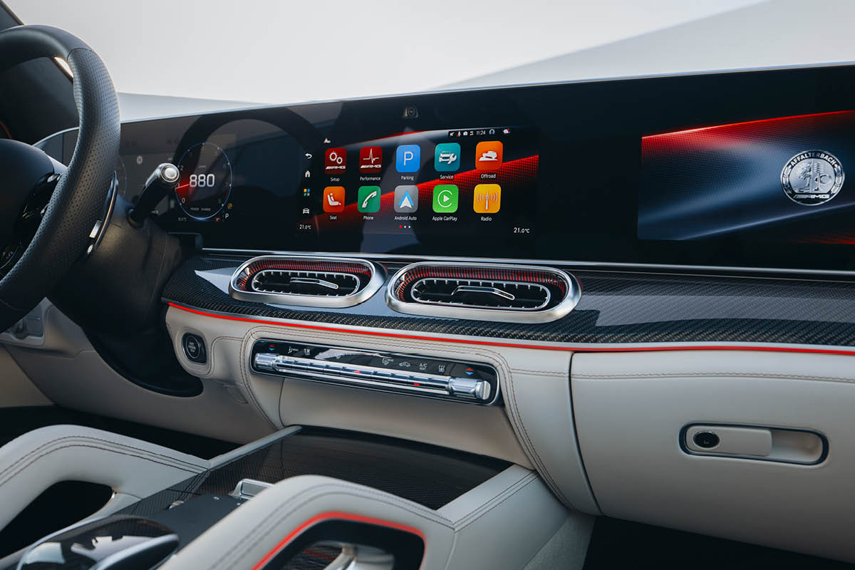 Dieses Bild zeigt den luxuriösen Innenraum des Fahrzeugs mit Fokus auf das Cockpit. Dominant ist ein großes, durchgehendes Display, das sowohl Instrumentenanzeige als auch Infotainment-System umfasst. Auf dem Bildschirm sind mehrere farbige App-Symbole sichtbar, darunter Navigation, Radio und Smartphone-Integration. Links befindet sich das digitale Kombiinstrument mit einer sportlichen Anzeige. Zwei große, runde Lüftungsdüsen mit metallischer Umrandung sitzen zentral im Armaturenbrett, das mit Carbon-Optik und feinen Ziernähten gestaltet ist. Eine rote Ambientebeleuchtung zieht sich dezent entlang der Armaturentafel und betont das hochwertige Design. Das Lenkrad ist teilweise sichtbar und ebenfalls sportlich ausgeführt.