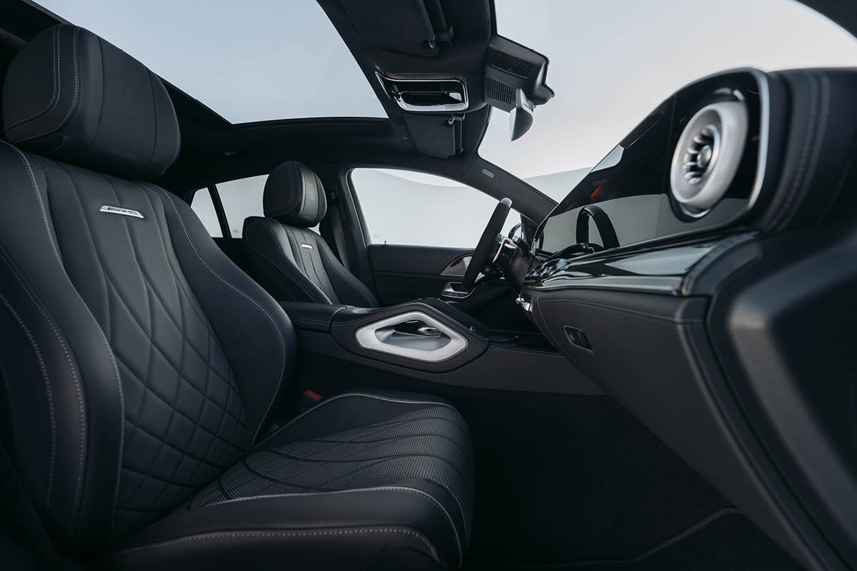 Das Bild zeigt den Innenraum des Fahrzeugs aus der Perspektive der Beifahrerseite mit Blick auf die beiden Vordersitze. Die Sitze sind in schwarzem Leder gehalten und aufwendig gesteppt, was einen hochwertigen und sportlichen Eindruck vermittelt. Auf den Sitzlehnen ist das AMG-Logo sichtbar. Die Mittelkonsole wirkt massiv und elegant mit einer silberfarbenen Einfassung und integrierten Bedienelementen. Das Armaturenbrett zieht sich klar und modern nach vorne, ergänzt durch runde Lüftungsdüsen im Turbinen-Design. Besonders auffällig ist das große Panoramaglasdach, das viel Licht in den Innenraum lässt und das Raumgefühl deutlich erweitert. Die Materialien wirken hochwertig, mit einer Kombination aus Leder, Metall und dunklen Zierelementen.