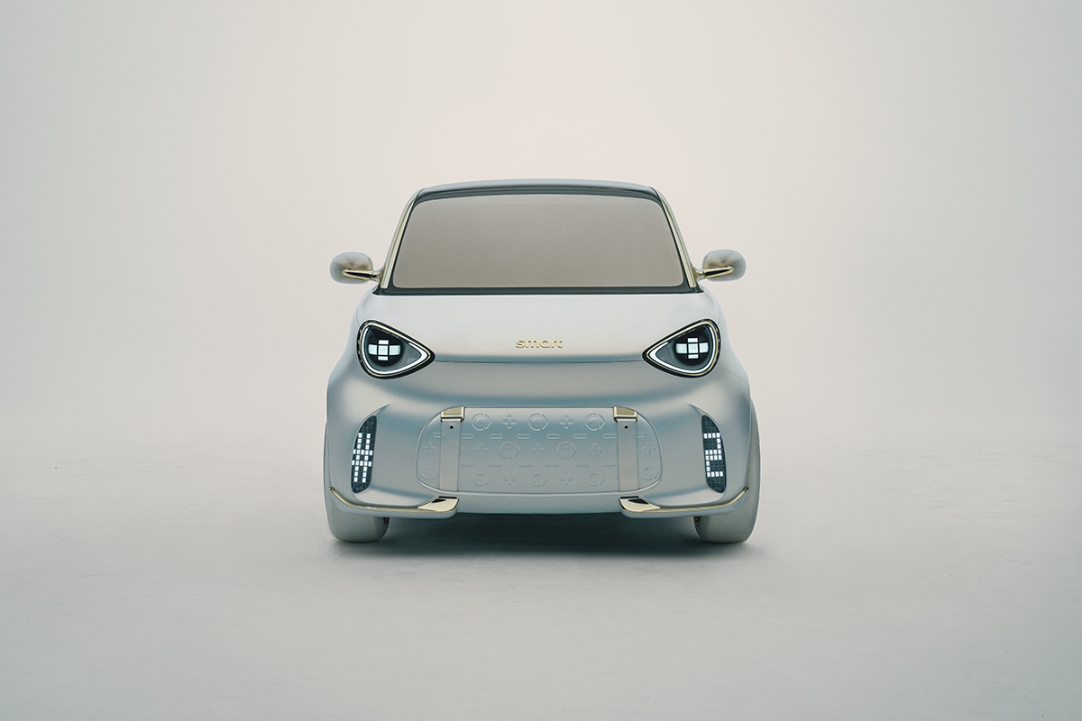 Frontansicht eines futuristischen weissen Smart Concept Autos mit markanten LED Scheinwerfern und goldenem Schriftzug. Die Frontpartie zeigt ein feines Muster und goldene Designelemente.
