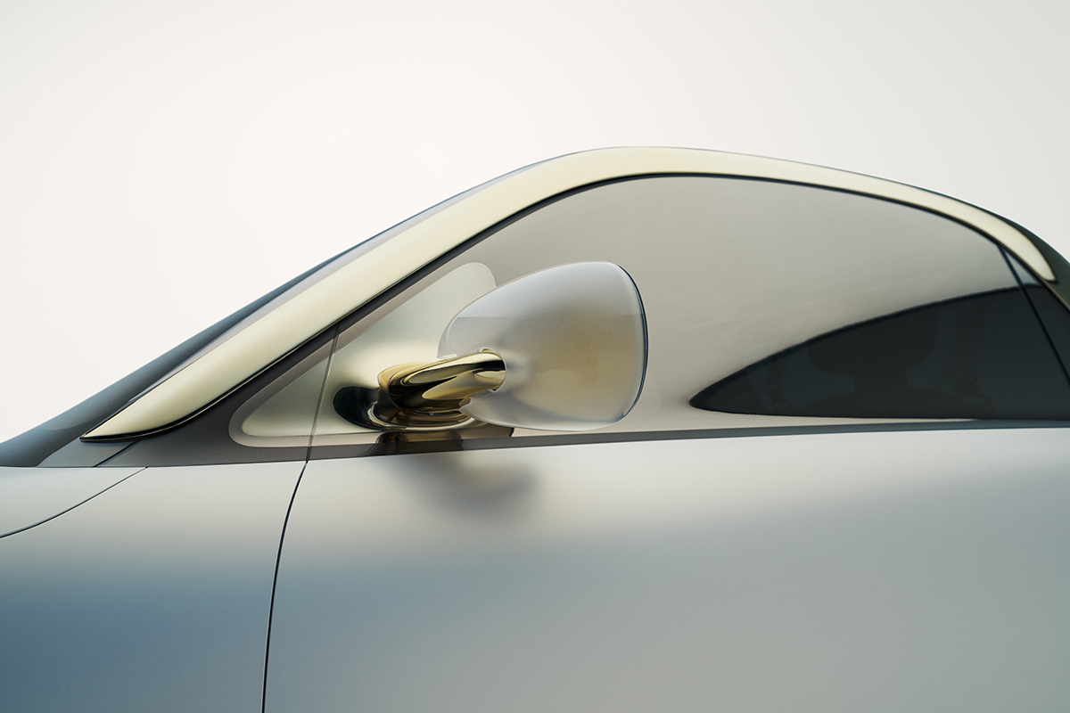 Detailaufnahme eines eleganten Aussenspiegels an einem weissen Smart Concept Auto. Der Spiegel hat ein halbtransparentes Gehaeuse und ist mit einer filigranen goldfarbenen Halterung befestigt.