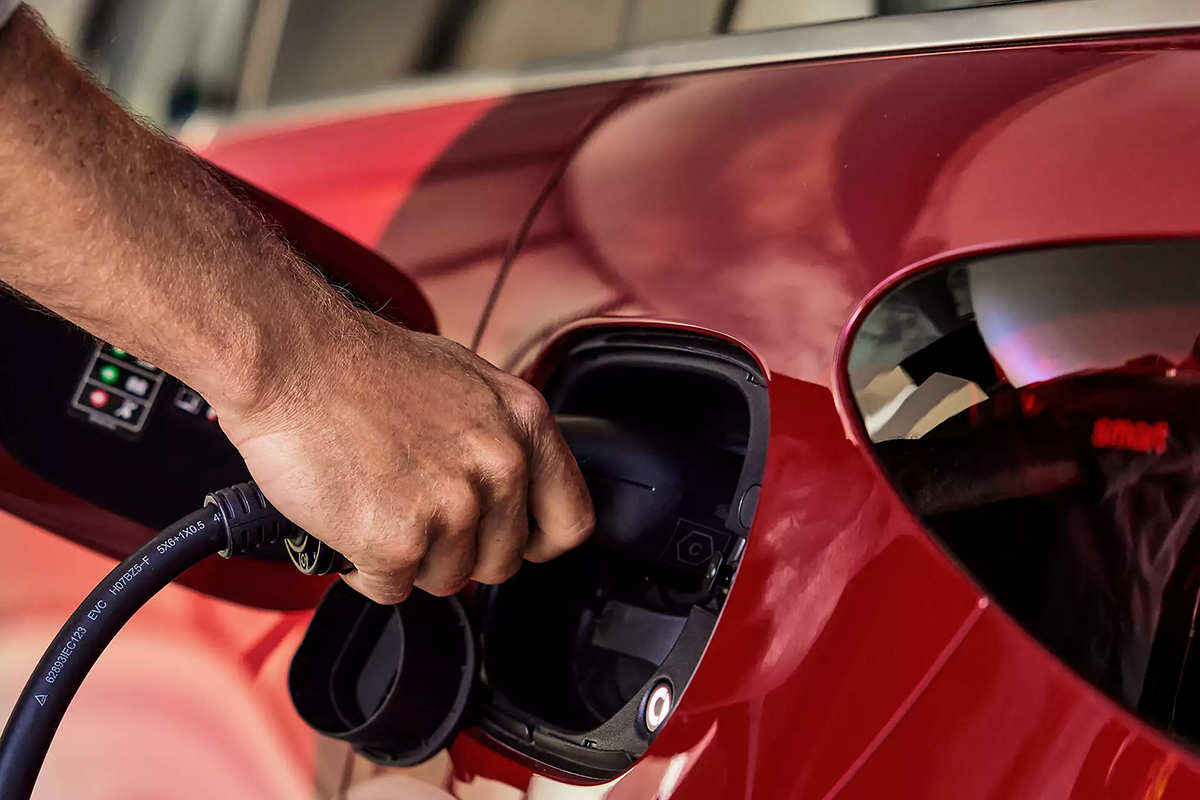 Das Bild zeigt eine Nahaufnahme eines roten Fahrzeugs, das gerade an eine Ladestation angeschlossen wird. Eine Hand hält den Stecker eines Ladekabels und führt ihn in die geöffnete Ladebuchse an der Fahrzeugseite ein. Der Bereich um die Ladebuchse ist schwarz umrandet. Ein Teil der glatten, glänzenden roten Karosserie ist sichtbar, ebenso ein seitliches Fahrzeugfenster, das spiegelnde Reflexionen zeigt. Im Hintergrund ist unscharf ein Teil einer Ladesäule zu erkennen, auf der ein kleines Display mit mehreren farbigen Symbolen oder Anzeigen leuchtet. Weitere Details oder Personen sind nicht im Bild.