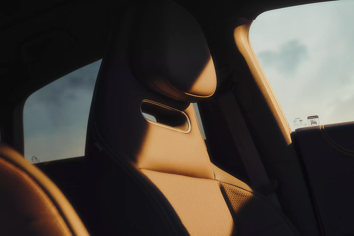 Das Bild zeigt eine Nahaufnahme des Innenraums eines Fahrzeugs. Im Mittelpunkt steht ein einzelner Sitz auf der vorderen Fahrzeugseite. Der Sitz ist aus einem glatten, hellbraunen Material gefertigt und verfügt über eine integrierte Kopfstütze. Unterhalb der Kopfstütze befindet sich eine ovale Öffnung mit einer glänzenden Umrandung. Die Sitzfläche und Teile der Lehne sind perforiert.