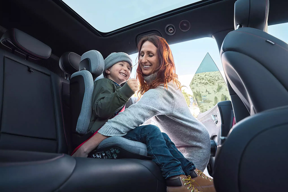 Das Bild zeigt den Innenraum eines Fahrzeugs aus der Perspektive der geöffneten Fondtür. Auf der Rückbank sitzt eine erwachsene Person, die sich leicht nach vorn beugt. Neben dieser Person sitzt ein Kind in einem Kindersitz, der seitlich erhöht und mit gepolsterten Elementen ausgestattet ist. Die erwachsene Person hält eine Hand am Kindersitz und ist in einer unterstützenden, stabilisierenden Position.