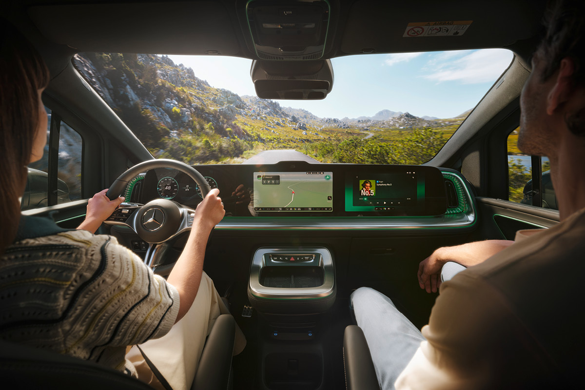 Blick aus dem Innenraum eines Mercedes-Benz auf eine kurvenreiche Straße in einer Berglandschaft. Das digitale Cockpit mit Navigations- und Infotainmentanzeige ist gut sichtbar, während zwei Personen im Fahrzeug sitzen.