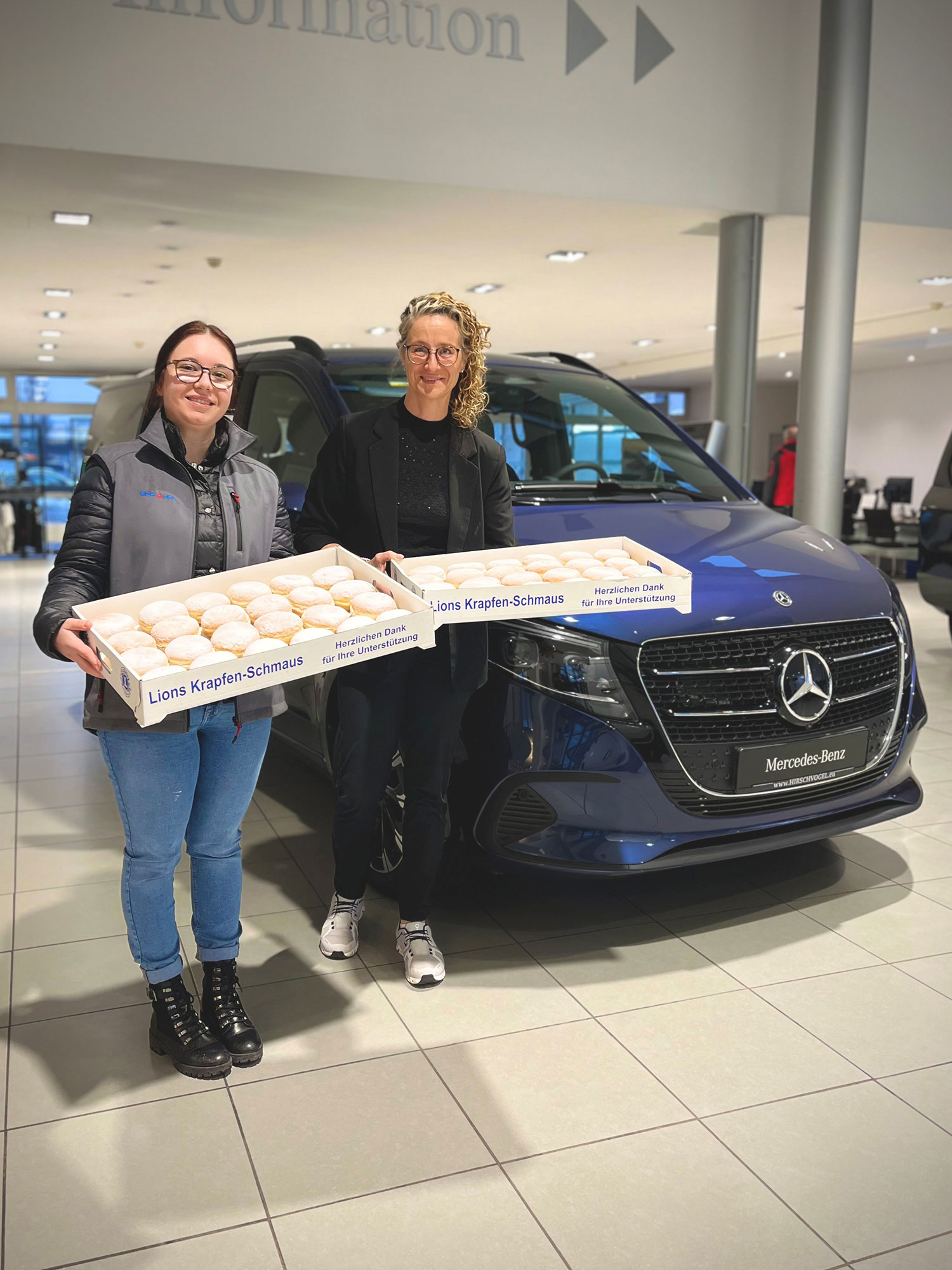 Auf dem Bild stehen zwei Personen in einem Autohaus vor einem dunkelblauen Mercedes‑Benz Fahrzeug. Beide halten jeweils eine große Kiste mit frisch gebackenen Krapfen, die mit „Lions Krapfen‑Schmaus“ beschriftet sind. Die Personen stehen seitlich vor dem Fahrzeug, das deutlich als Transporter erkennbar ist. Im Hintergrund sind die hell ausgeleuchteten Verkaufsräume des Autohauses mit weiteren Fahrzeugen und Beratungstischen zu sehen.