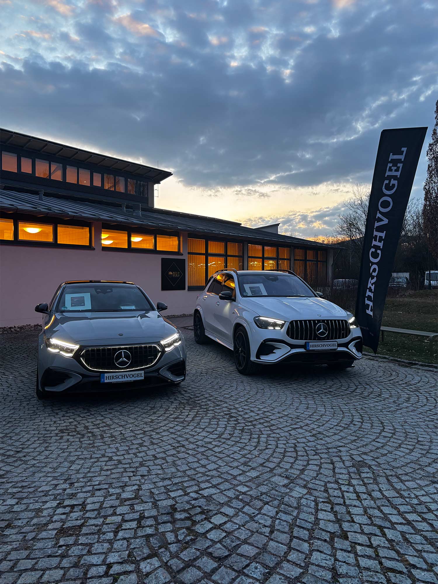 Zwei Mercedes Fahrzeuge stehen auf gepflastertem Platz vor einem Gebaeude bei Abenddaemmerung, links ein graues Modell, rechts ein weisses SUV, daneben eine schwarze Hirschvogel Fahne.