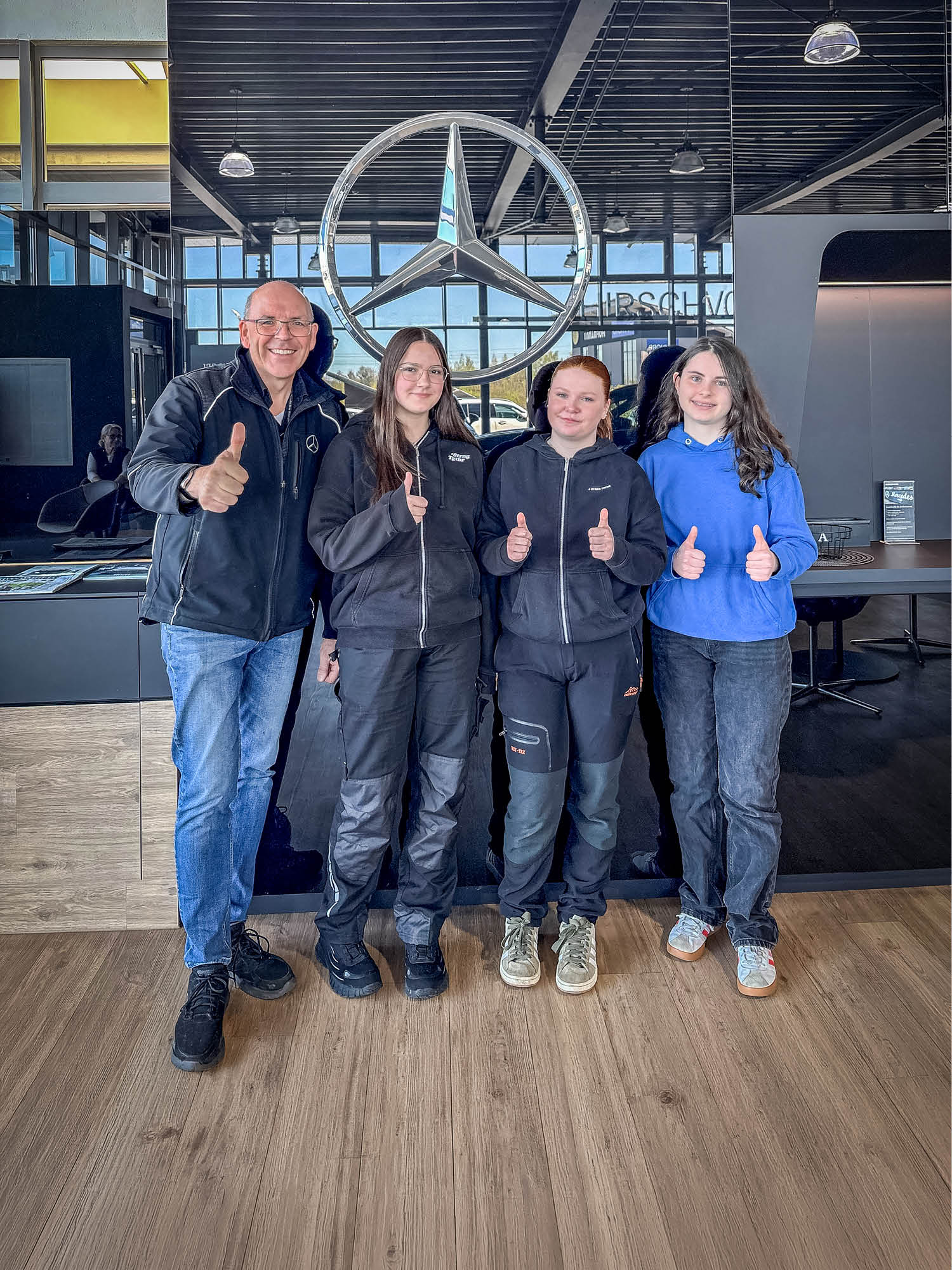 Girlsday 2026 - Auf dem Bild sind vier Personen zu sehen, die nebeneinander in einem modernen Innenraum stehen. Der Raum wirkt wie ein Autohaus oder ein Ausstellungsbereich, was durch das große, silberne Mercedes‑Benz‑Logo an der Wand im Hintergrund deutlich wird. Die Umgebung ist hell und offen gestaltet, mit großen Fensterflächen, dunklen Möbeln und einer Decke aus Metallstreben mit mehreren Hängeleuchten. Der Boden besteht aus hellem Holz oder Holzimitat. Alle vier Personen stehen frontal zur Kamera und heben jeweils einen Daumen nach oben. Die Kleidung wirkt überwiegend praktisch und funktional: Drei der Personen tragen dunkle Arbeits- oder Freizeitjacken mit passenden Hosen, eine Person trägt einen blauen Kapuzenpullover zu dunklen Jeans. Die Schuhe sind sportlich beziehungsweise robust. Hinter den Personen befindet sich ein schwarzer Tresen oder Tisch mit Informationsmaterial. Durch die Glasflächen im Hintergrund ist Tageslicht sichtbar, wodurch der Raum freundlich und klar beleuchtet erscheint. Die Bildkomposition ist symmetrisch, wobei das große Markenlogo zentral über den Personen angebracht ist und als auffälliger Blickfang dient.