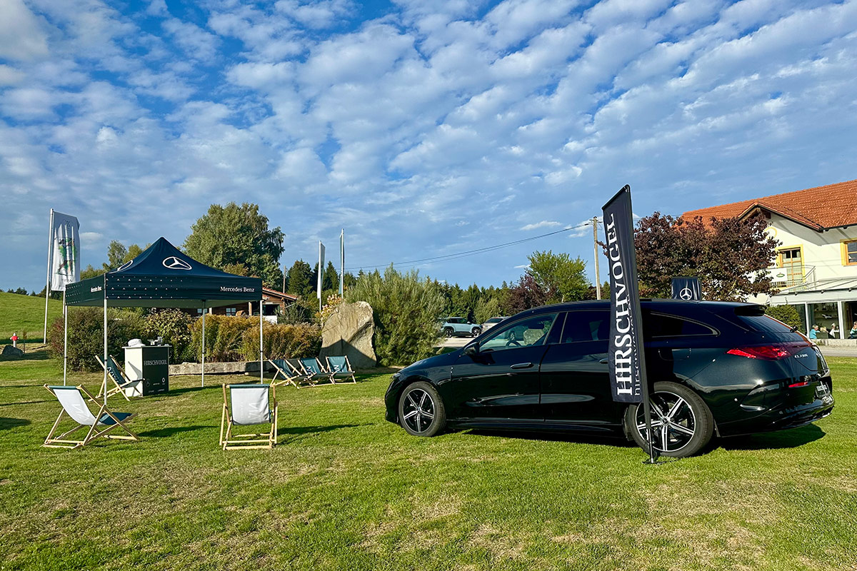 Auf einer Rasenfläche eines Golfplatzes ist ein schwarzes Mercedes-Benz Fahrzeug ausgestellt. Daneben befindet sich eine schwarze Fahne mit dem Schriftzug „HIRSCHVOGEL“. Links im Bild steht ein schwarzes Mercedes-Benz Eventzelt mit Mercedes-Benz Logo. Vor dem Zelt sowie auf der Grünfläche sind mehrere Liegestühle aufgestellt.
