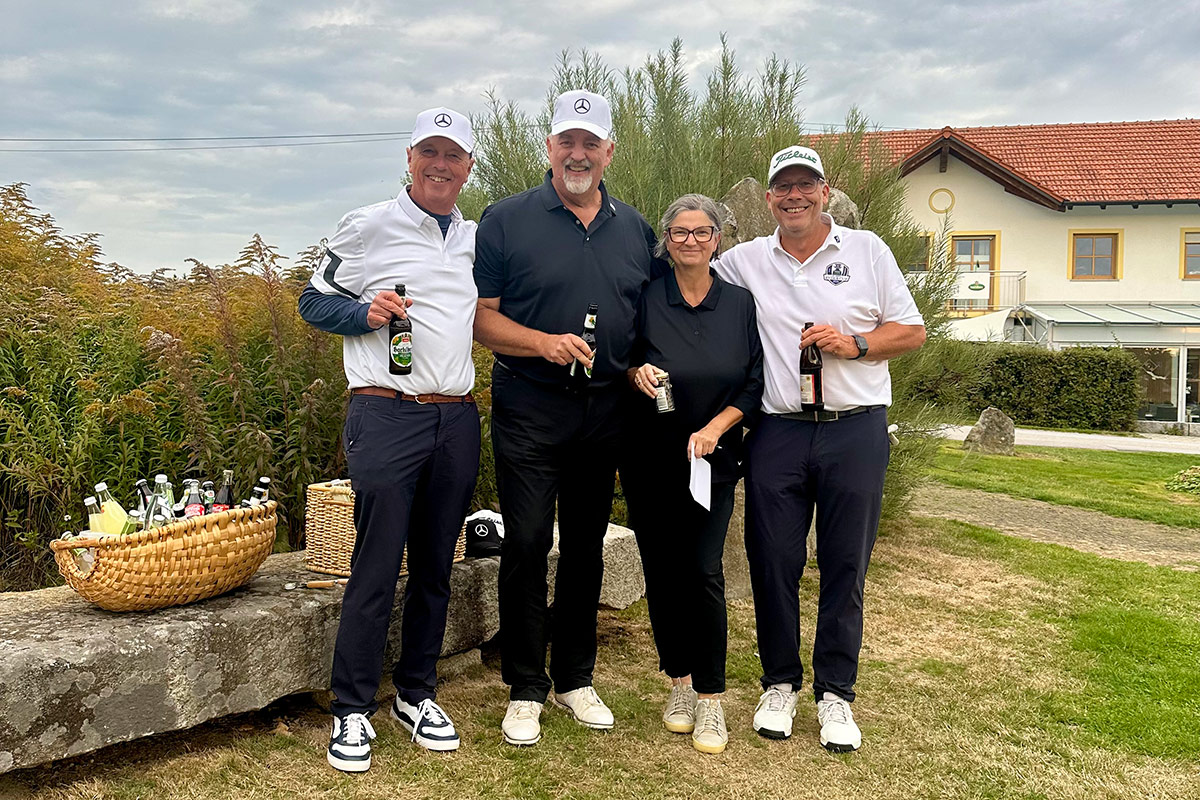 Vier Personen in Freizeit- und Golfkleidung stehen für ein Gruppenfoto auf einer Rasenfläche eines Golfclubs. Die Personen halten jeweils Getränkeflaschen oder ein Dokument in den Händen. Links im Bild befindet sich auf einer Steinbank ein großer Korb mit verschiedenen Getränkeflaschen.