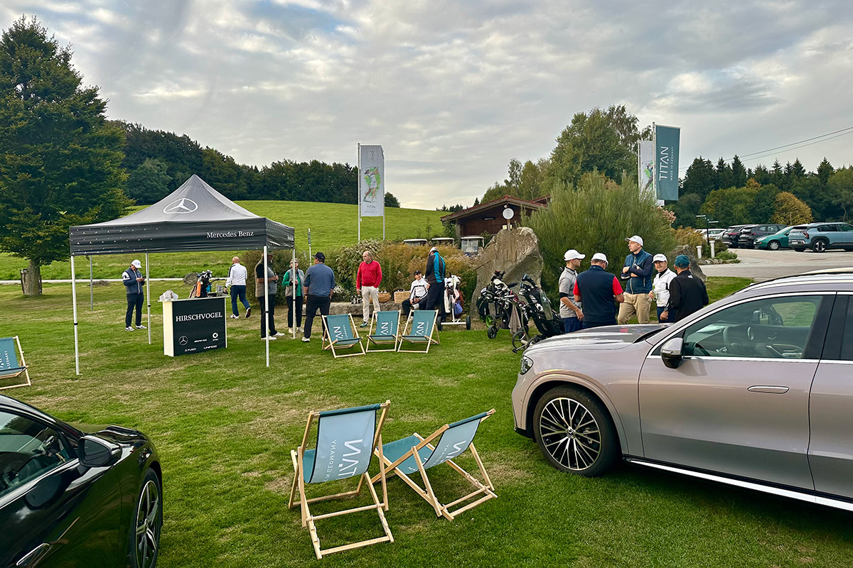 Auf einer gepflegten Rasenfläche eines Golfclubs findet eine Veranstaltung im Freien statt. Mehrere Personen stehen in kleinen Gruppen zusammen und unterhalten sich neben Golfbags und Golftrolleys. Links befindet sich ein schwarzes Eventzelt mit Mercedes-Benz Logo und der Aufschrift „Mercedes-Benz“. Vor dem Zelt steht eine Informationssäule mit dem Schriftzug „HIRSCHVOGEL“.