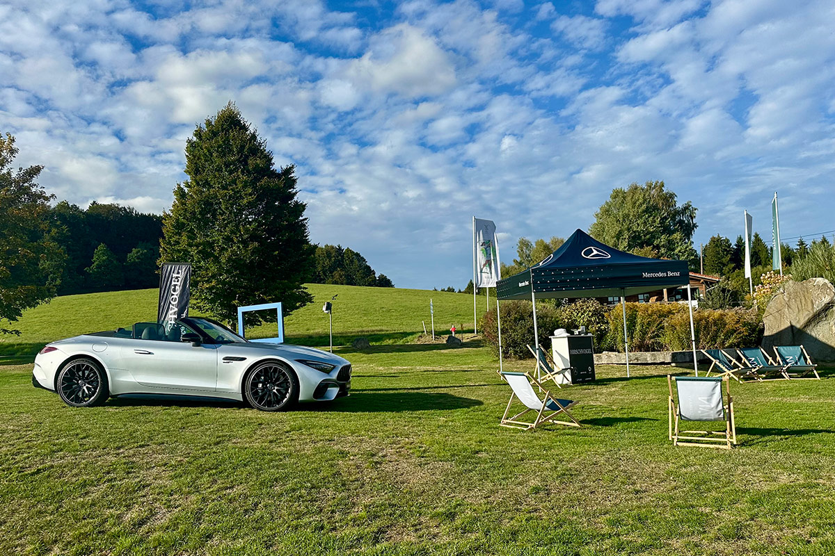 Auf einer weitläufigen Rasenfläche eines Golfplatzes steht ein weißes Mercedes-Benz Cabriolet mit geöffnetem Verdeck. Rechts daneben befindet sich ein schwarzes Mercedes-Benz Eventzelt mit Mercedes-Benz Logo. Vor und neben dem Zelt sind mehrere Liegestühle aufgestellt. Im Umfeld stehen Fahnen von Hirschvogel und Mercedes-Benz.