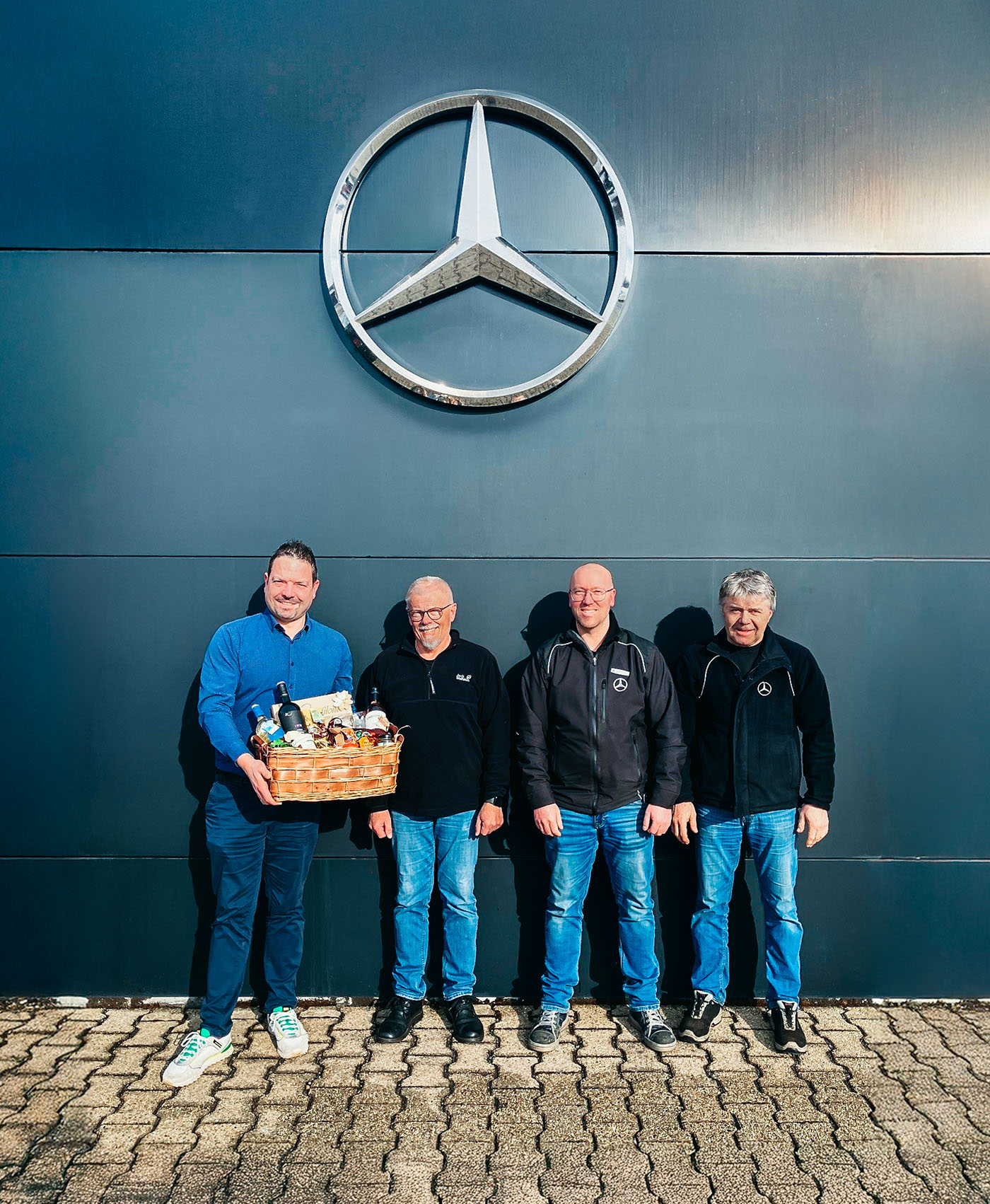 Vier Personen stehen vor einen Wand, lachen in die Kamera und überreichen einen Presentkorb. Zu sehen ist das Mercedes Logo über Ihnen.