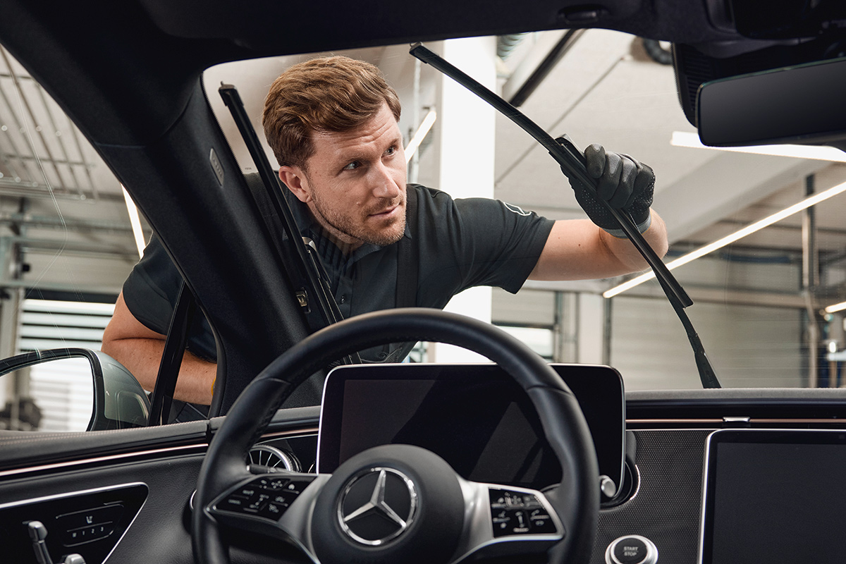 Das Bild zeigt die Innenansicht eines geparkten Autos. Im Vordergrund ist das Lenkrad mit dem Mercedes‑Benz‑Logo sowie das zentrale Display sichtbar. Durch die Frontscheibe erkennt man eine Person, die außen am Fahrzeug steht und ein Wischerblatt austauscht. Die Person trägt schwarze Handschuhe und hält den Wischerarm mit einer Hand fest, während sie mit der anderen das neue Wischerblatt montiert. Im Hintergrund ist eine helle Werkstattumgebung mit Metallstrukturen und Beleuchtung zu sehen.